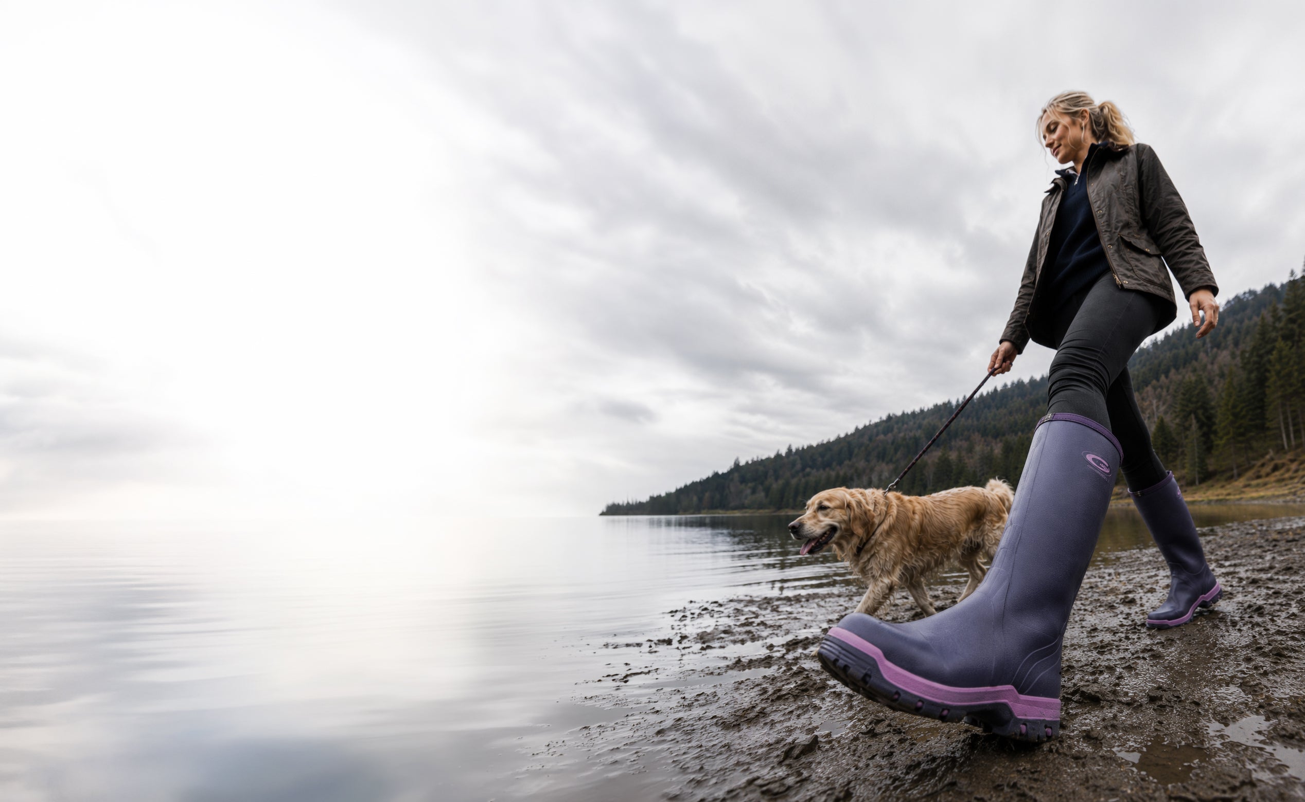 A woman walks her dog along a muddy lakeshore under an overcast sky, wearing Grubs Rainline 2.0 The water is calm, with a forested shoreline in the distance.